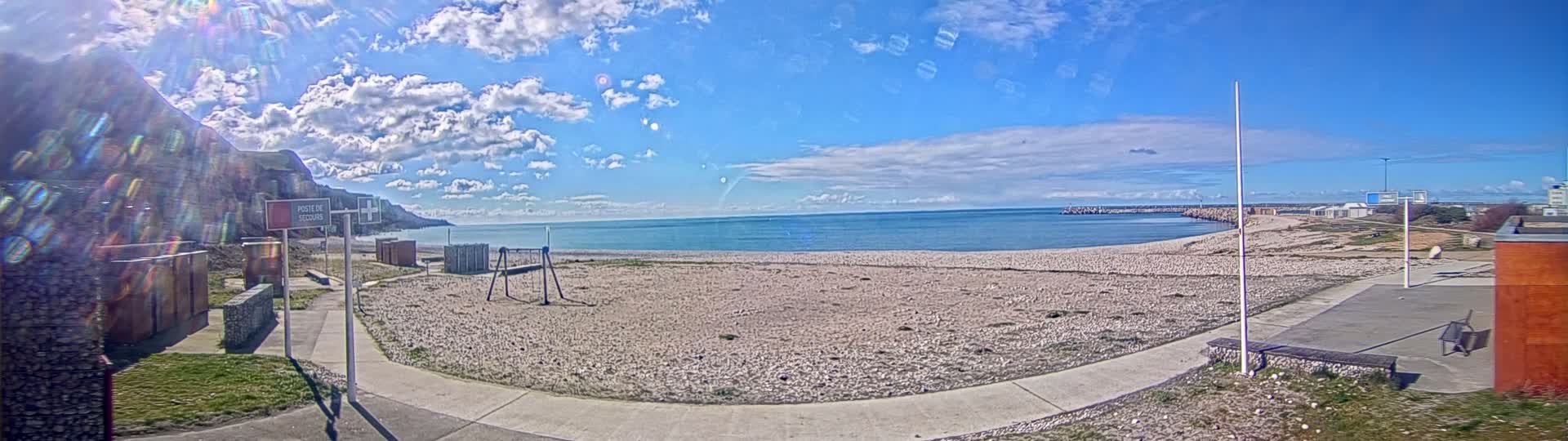 A mostly sunny day at a beach with a calm sea, a rocky shore, and a playground on the sand.