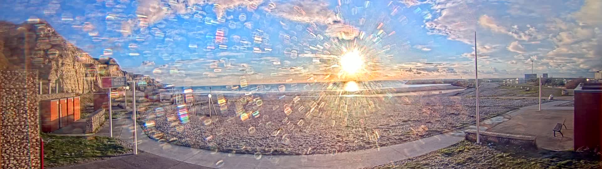 A bright sun sets over a sandy beach with a stone cliff in the background under a partly cloudy sky.