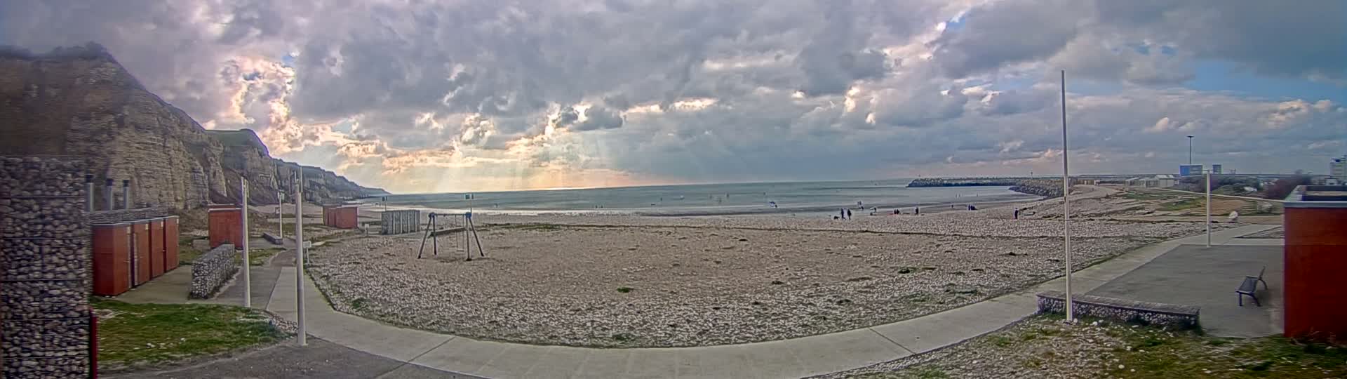 A mostly cloudy day reveals a pebble beach with a few people, a children's play area, and changing rooms situated beneath a steep cliff face next to a calm sea.