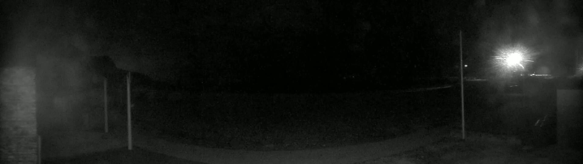 A dark, nighttime scene shows a paved area with two thin poles and a bright light source in the distance.