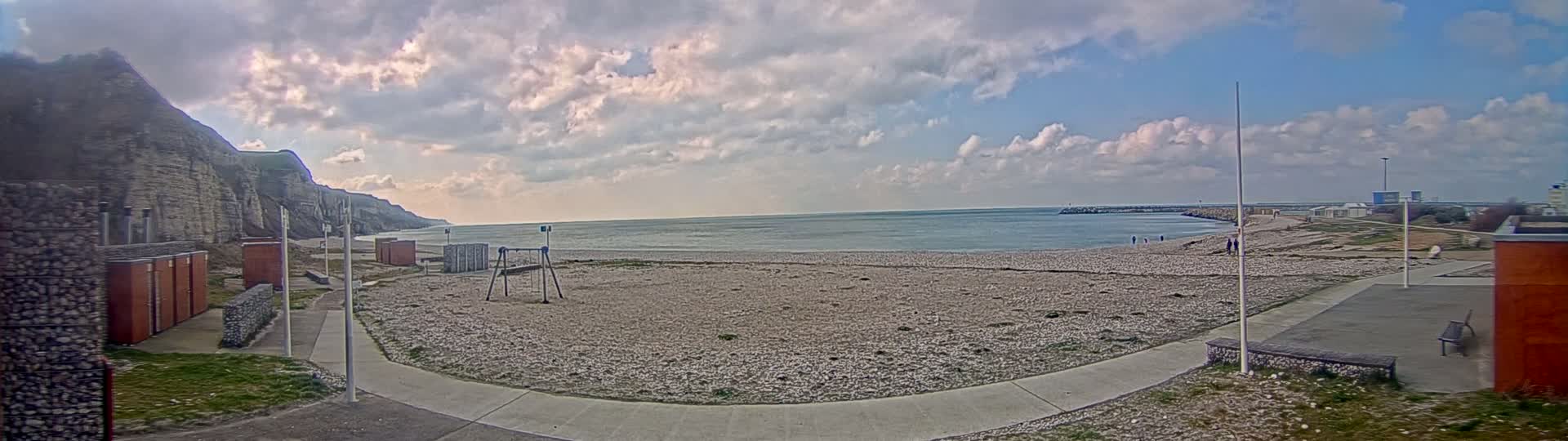 A mostly cloudy day reveals a pebble beach with a few people walking, a playground, and restroom facilities situated between a calm sea and a steep cliff.
