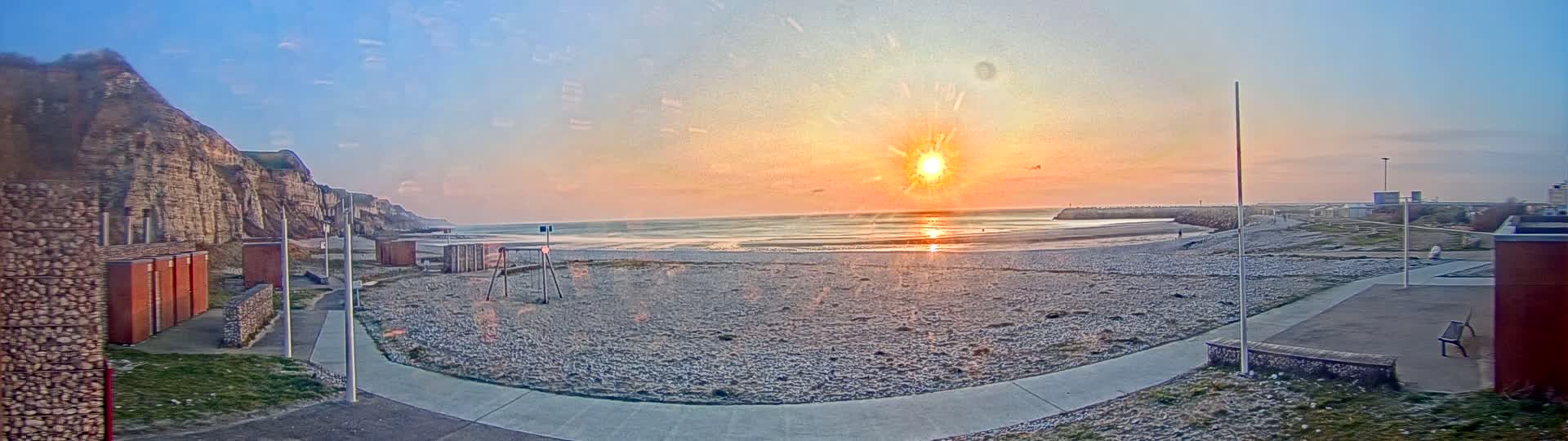A beach at sunset, with a pebble shore, cliffs in the background, and a children's play area nearby.
