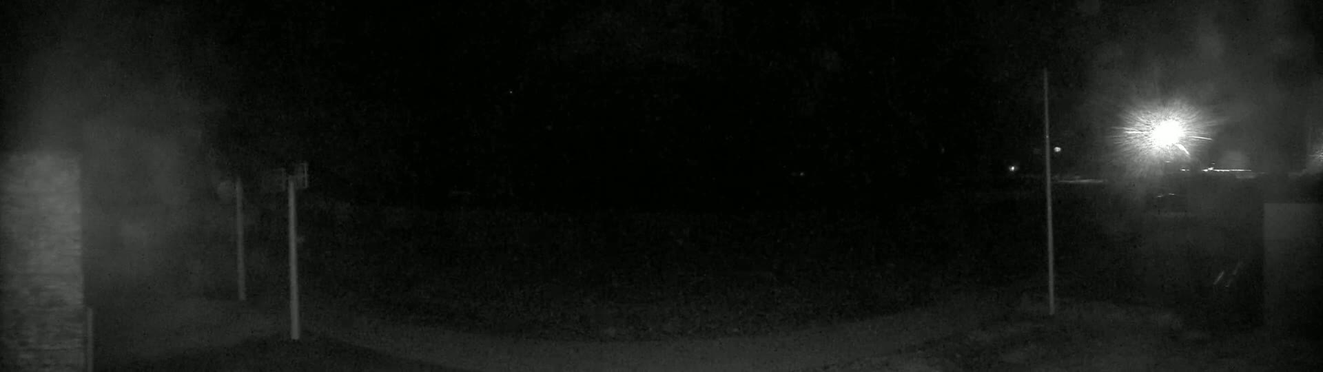A dark, nighttime scene shows a dimly lit area with two vertical poles and a bright light source in the upper right corner, possibly under snowy conditions.