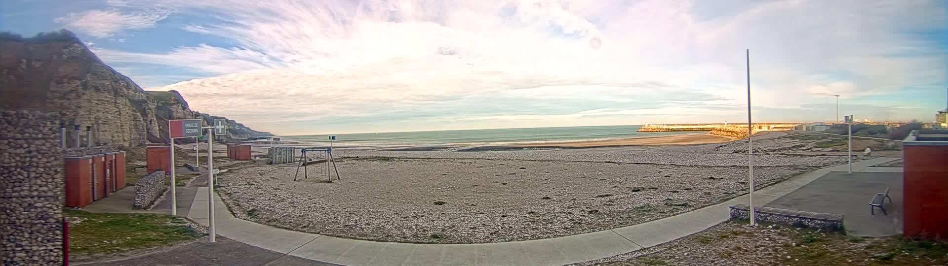 A mostly sunny day at a pebble beach with a rocky cliff to the left, a calm sea in the distance, and a playground visible on the beach.