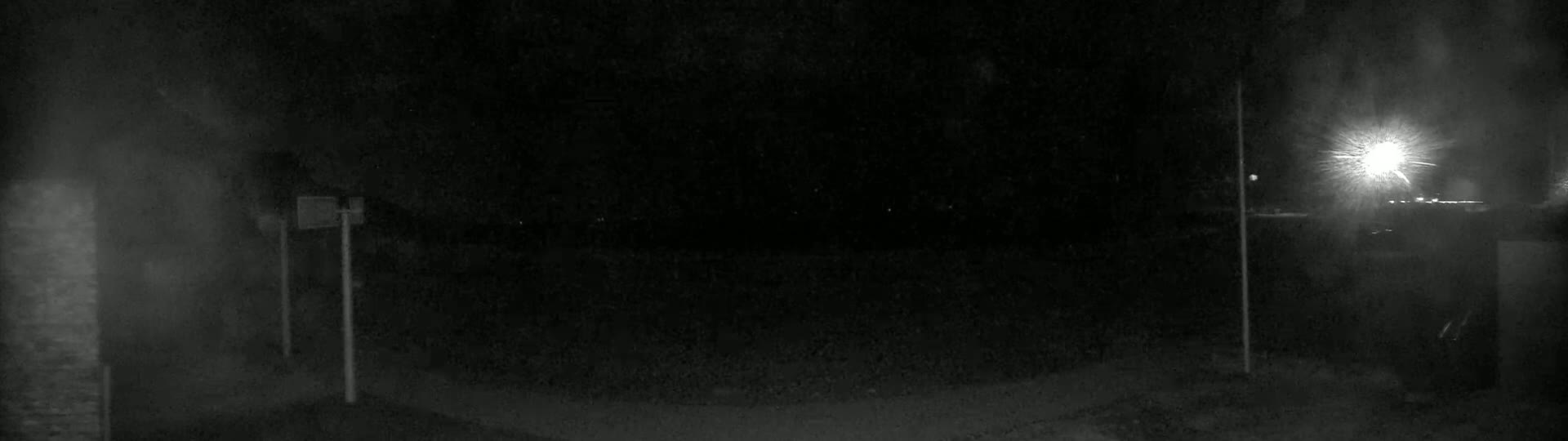 A nighttime, low-light outdoor scene shows a dark, mostly empty area with two tall, thin poles and a bright light source in the distance.