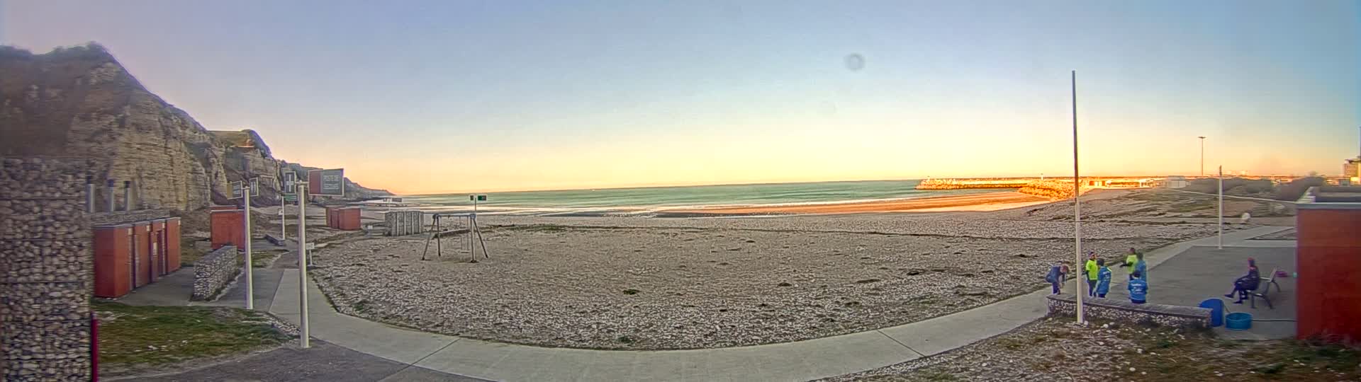 A sandy beach stretches out to a calm ocean under a clear, sunset sky, with a rocky cliff face to the left and several people gathered near a walkway on the right.