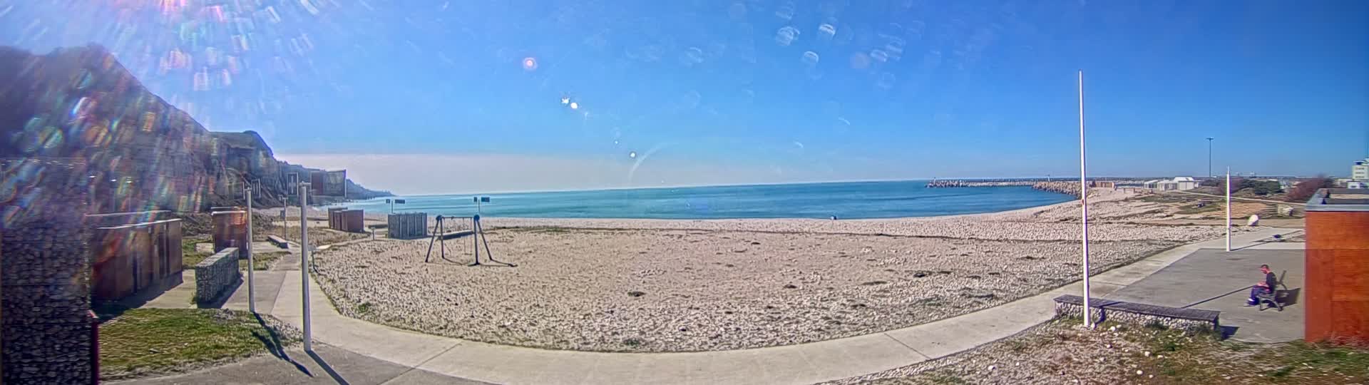 A sandy beach with a calm blue sea stretches out under a clear, sunny sky, backed by a rocky cliff and a small harbor.