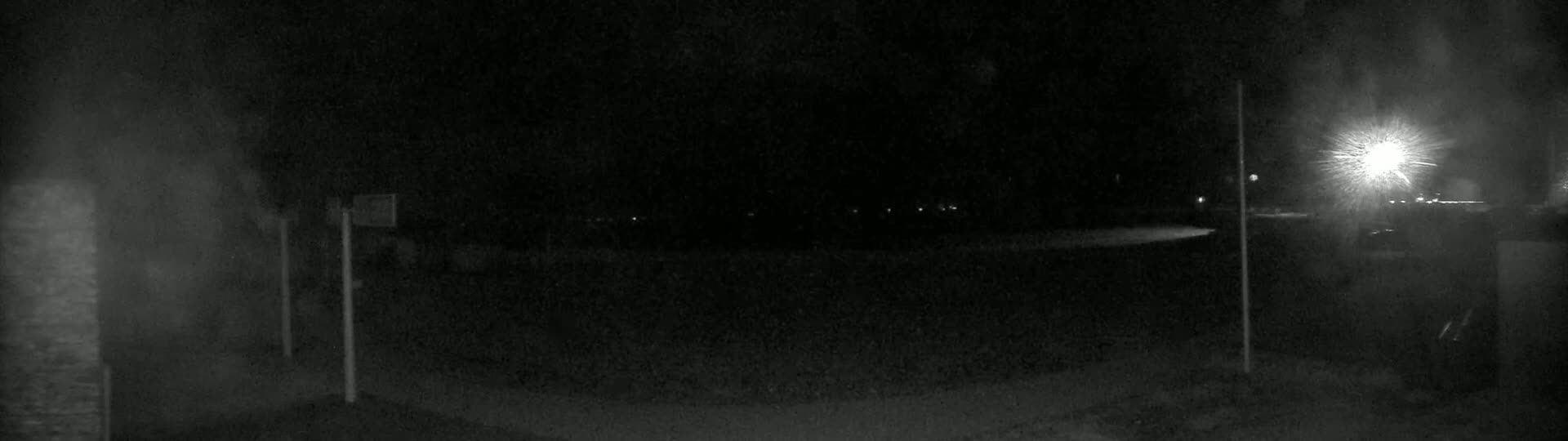 A nighttime, low-light view of a dark, mostly empty paved area with a bright light source in the distance, and some indistinct objects or structures in the foreground.