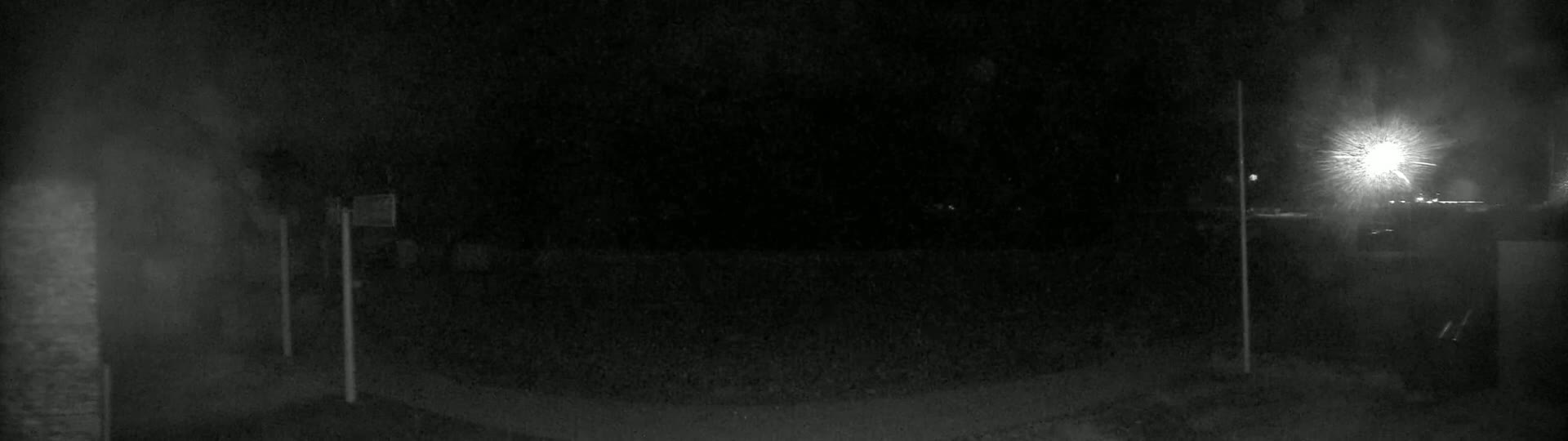 A nighttime, low-light image shows a dark, empty area with two poles and a bright light source in the distance.