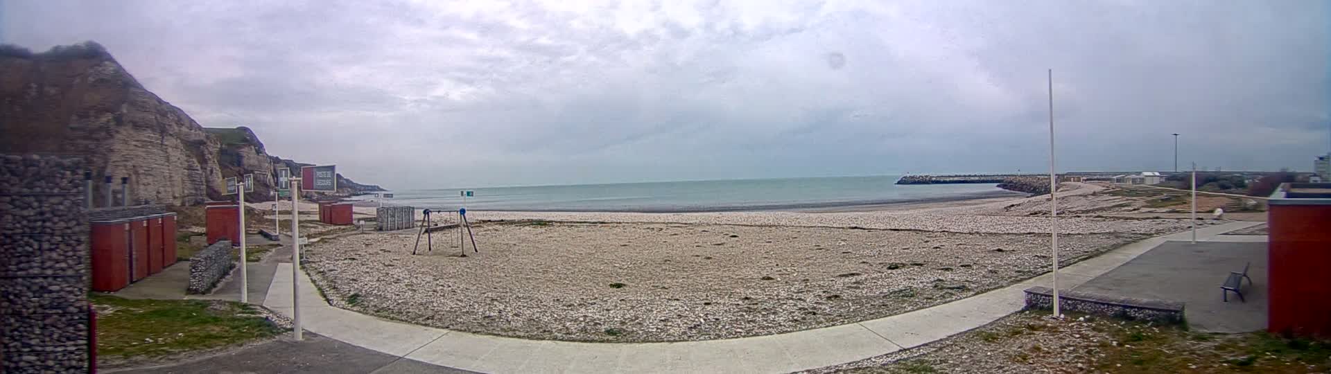 A pebble beach stretches out to a calm sea under an overcast sky, backed by white cliffs and small buildings.