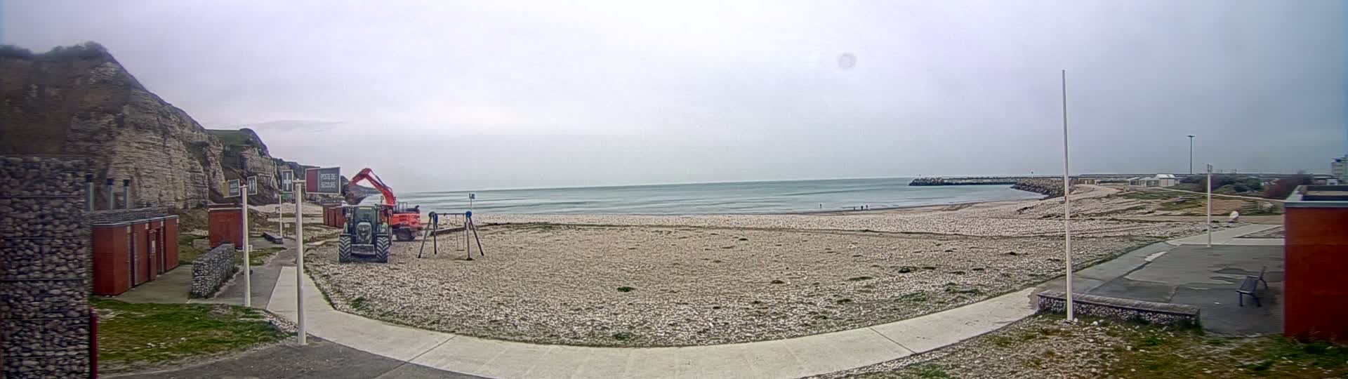 A mostly cloudy day at a beach shows construction equipment near a children's playground, a calm sea, and a cliff in the background.