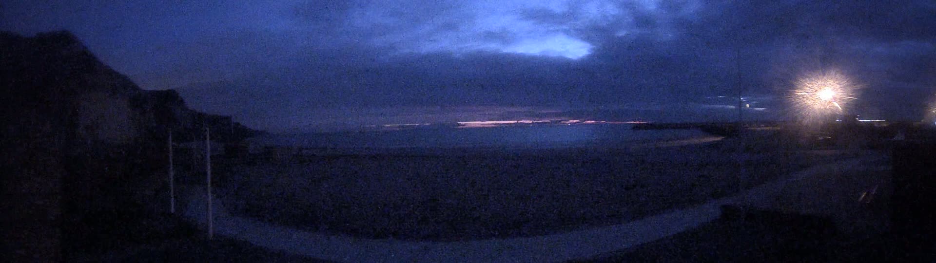 A dark, nighttime scene shows a body of water under a cloudy sky, with a bright light emanating from a source on the right, and a dark landmass to the left.