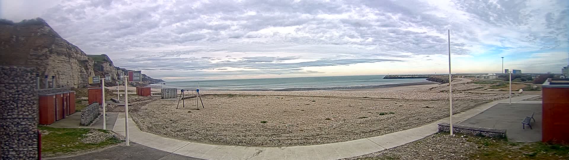 A mostly cloudy day reveals a beach with a playground, small buildings, and a walkway leading to the ocean, backed by a cliff face.