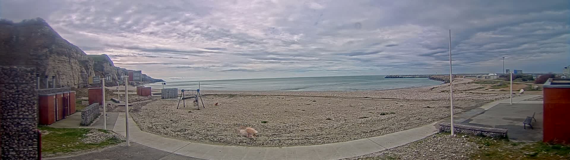 A mostly cloudy day reveals a sandy beach with a playground, a small harbor, and a cliff in the background.