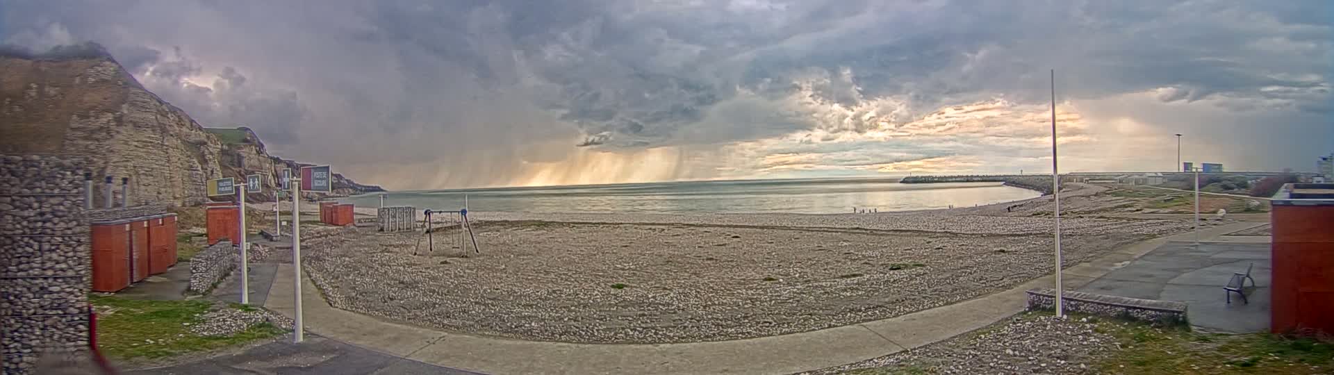 A pebble beach stretches out to a calm sea under a dramatic sky filled with dark rain clouds, with a portion of the sky showing a lighter, brighter section.