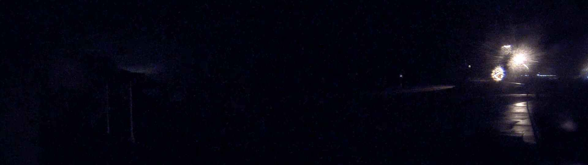 A nighttime, dimly lit outdoor scene shows a wet paved area with bright lights reflecting in the water.