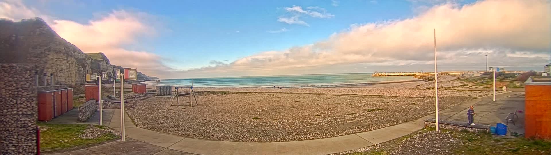 A sandy beach with a calm ocean stretches out under a partly cloudy sky with a pinkish hue near the horizon, backed by a cliff and small buildings.