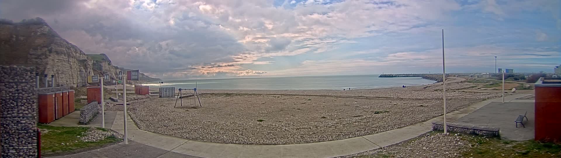 A mostly cloudy day shows a pebble beach with a playground, a walkway, and white cliffs in the background, next to a calm sea with a breakwater.