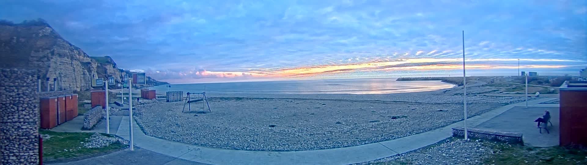 A pebble beach at sunrise, under a mostly clear sky with streaks of pink and orange, is bordered by a cliff face and some buildings, and features a small playground.