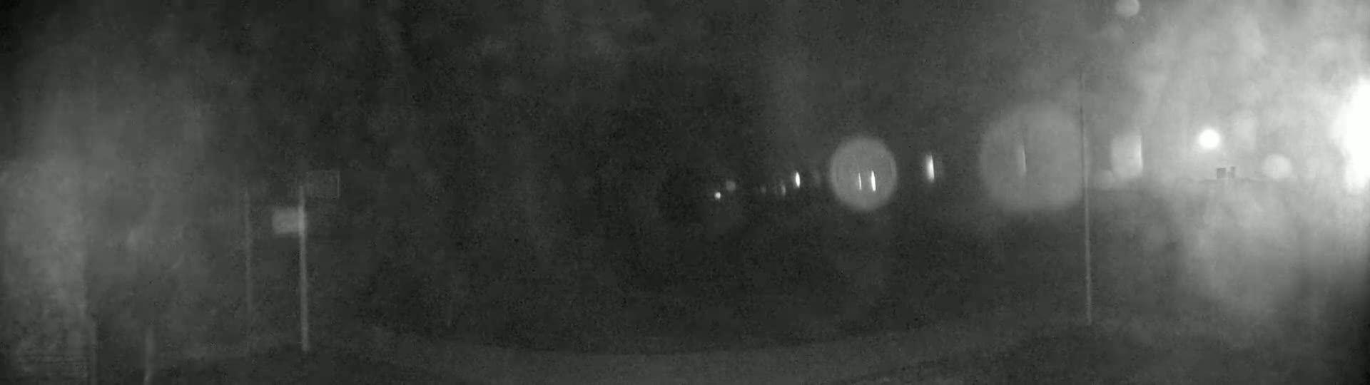 A blurry, nighttime image shows a roadway with indistinct objects and out-of-focus lights in foggy conditions.