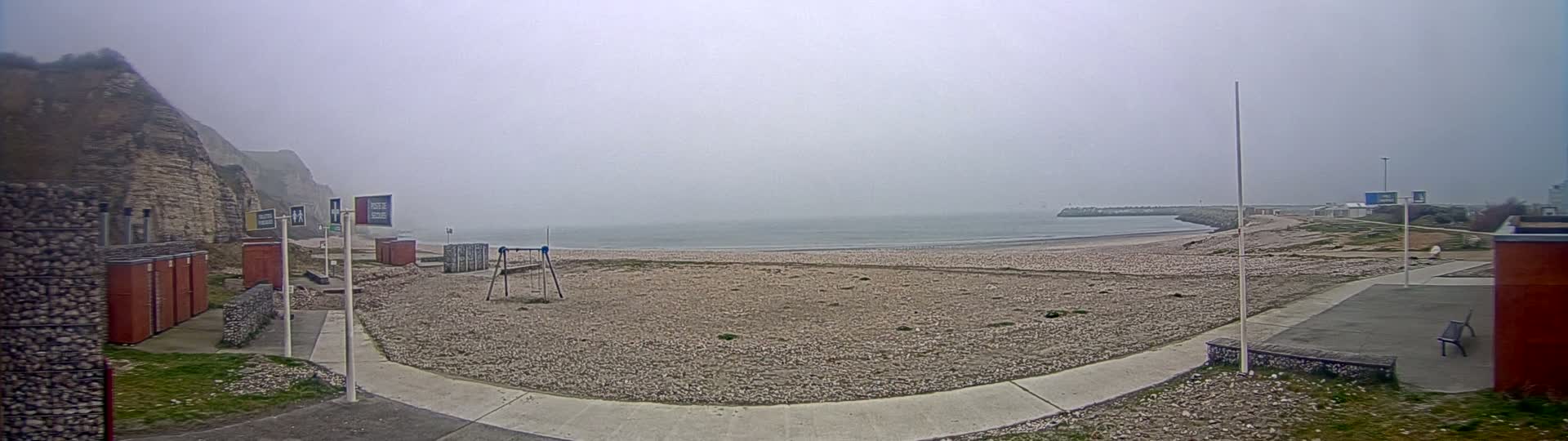 A foggy beach with a pebble shore, a children's swing set, and a stone cliff in the background.