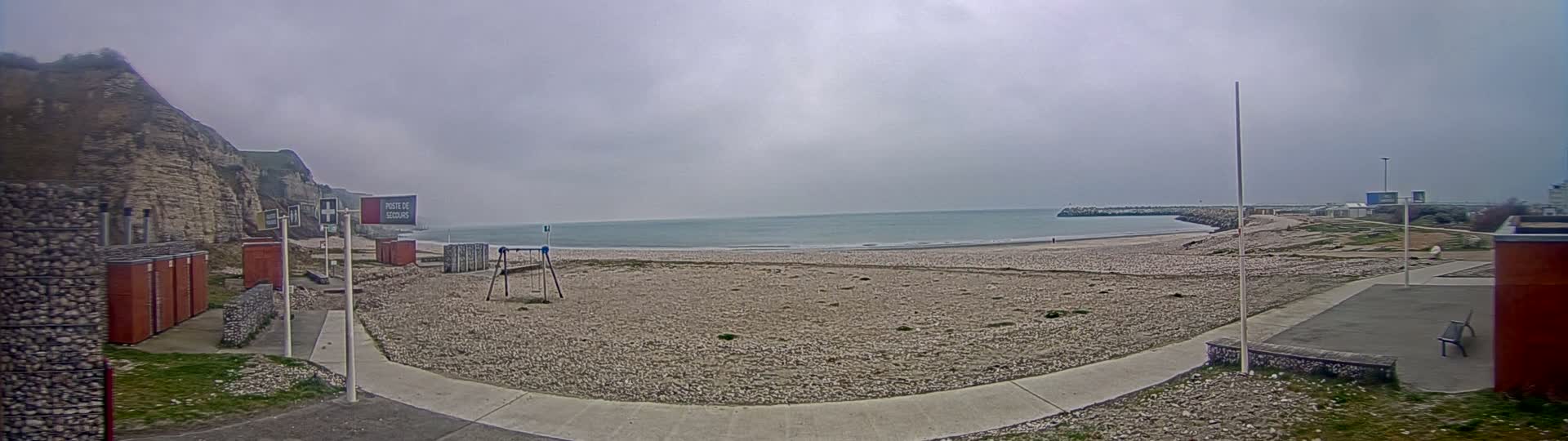 A mostly empty pebble beach stretches out to a calm, gray sea under an overcast sky, backed by a cliff face and a small playground.