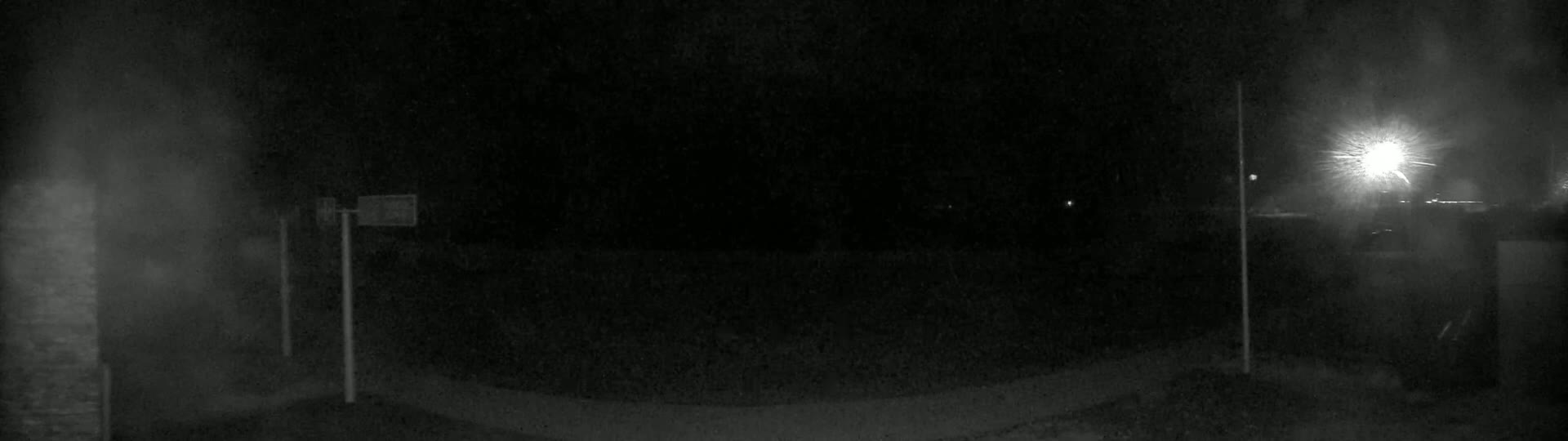 A dark, nighttime scene shows a paved area with two tall poles and a bright light in the distance, partially obscured by what appears to be fog or dust.