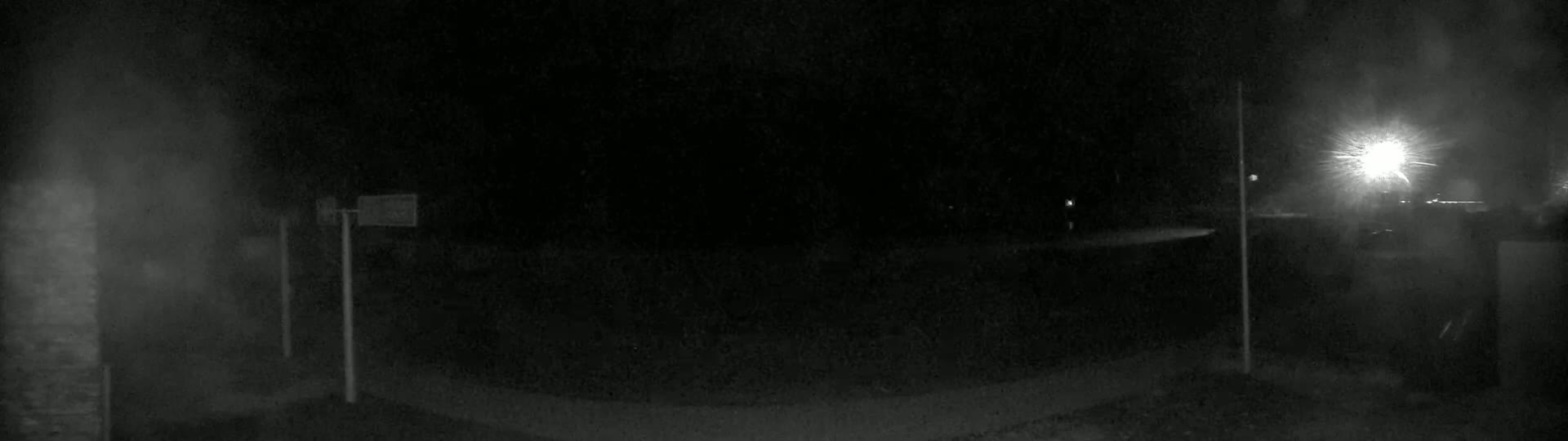 A dark, nighttime scene shows a dimly lit road with two vertical poles and a bright light source in the distance.