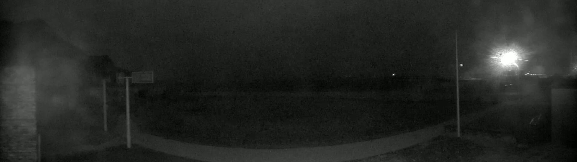 A nighttime, low-light image shows a dark, mostly empty paved area with indistinct structures and a bright light source in the distance.