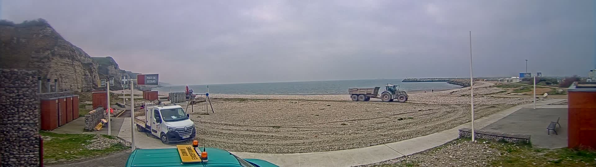A tractor and trailer are parked on a beach next to a cliff face under an overcast sky, with a van and other vehicles nearby.