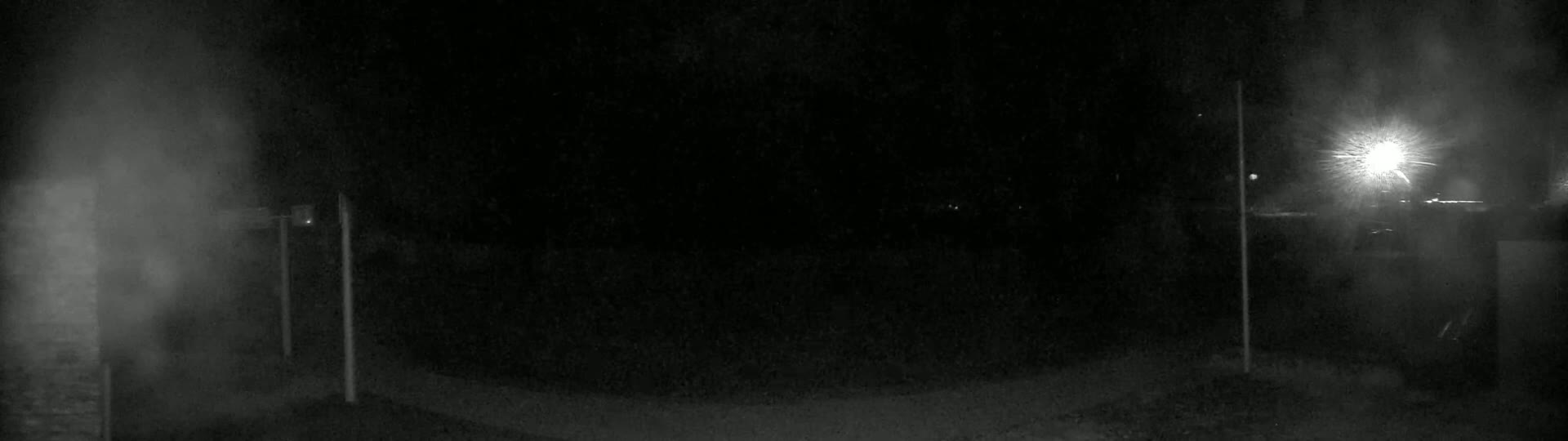 A nighttime, dimly lit outdoor scene shows a dark, empty area with two vertical poles and a bright light source in the distance.