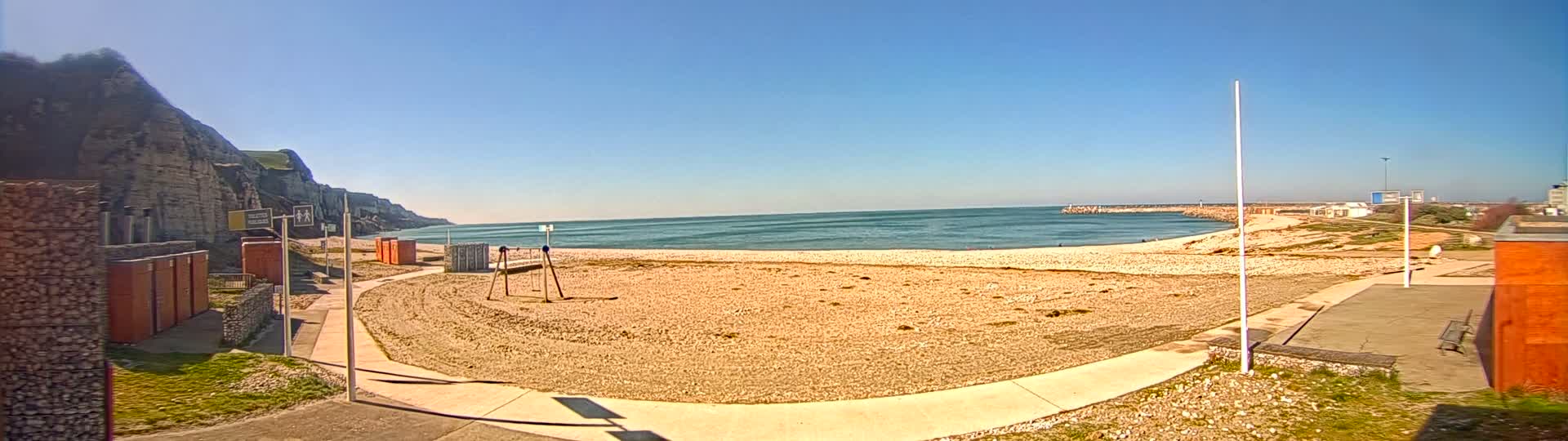 A sandy beach stretches out to a calm, blue ocean under a clear, sunny sky, backed by a rocky cliff face and a paved walkway.