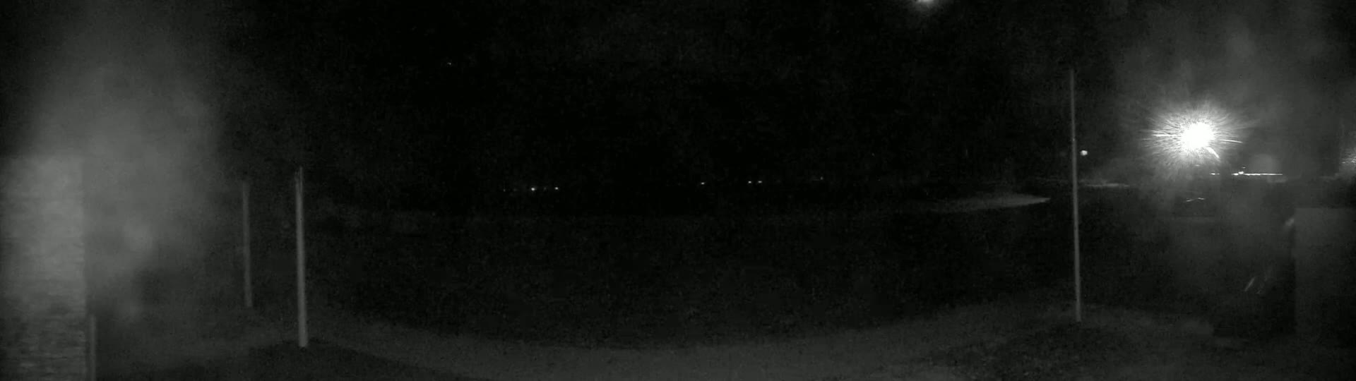 A dark, nighttime scene shows a dimly lit area with some faint lights in the distance and two tall, thin poles standing on either side of the frame, under a possibly snowy or hazy atmosphere.
