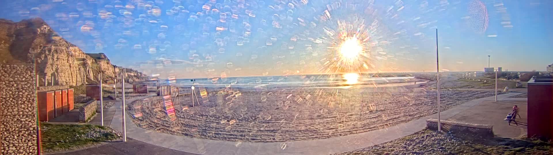 A beach scene with a bright, sun-starburst effect, showing sandy beach, ocean waves, and a cliff in the background under a mostly clear sky.