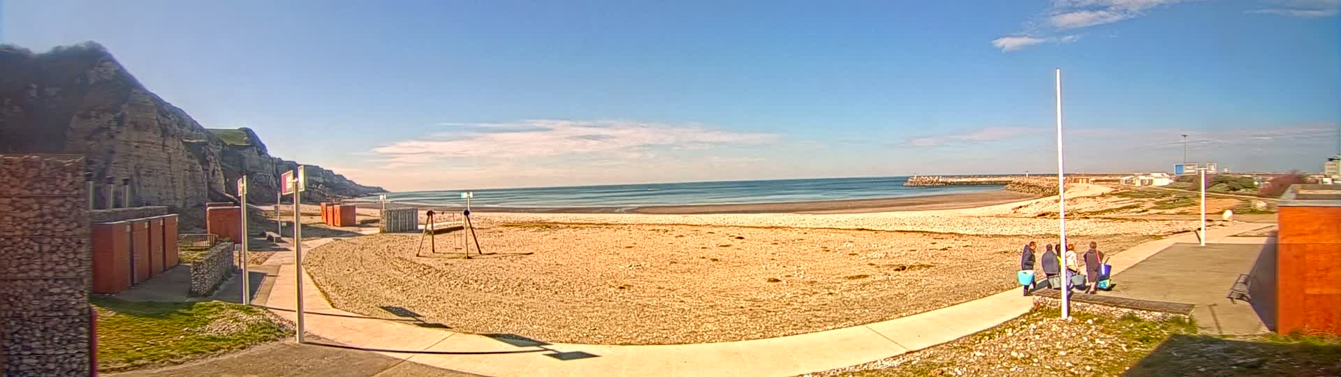 A sandy beach with a few people and buildings is visible under a mostly sunny sky near a rocky cliff.