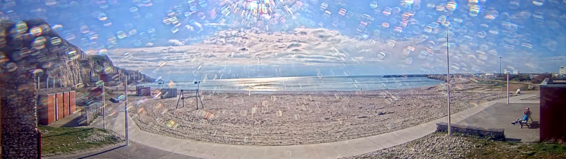 A mostly sunny day at a sandy beach with a calm sea, partially obscured by a shimmering effect, shows a paved walkway, buildings, and a person sitting on a bench.