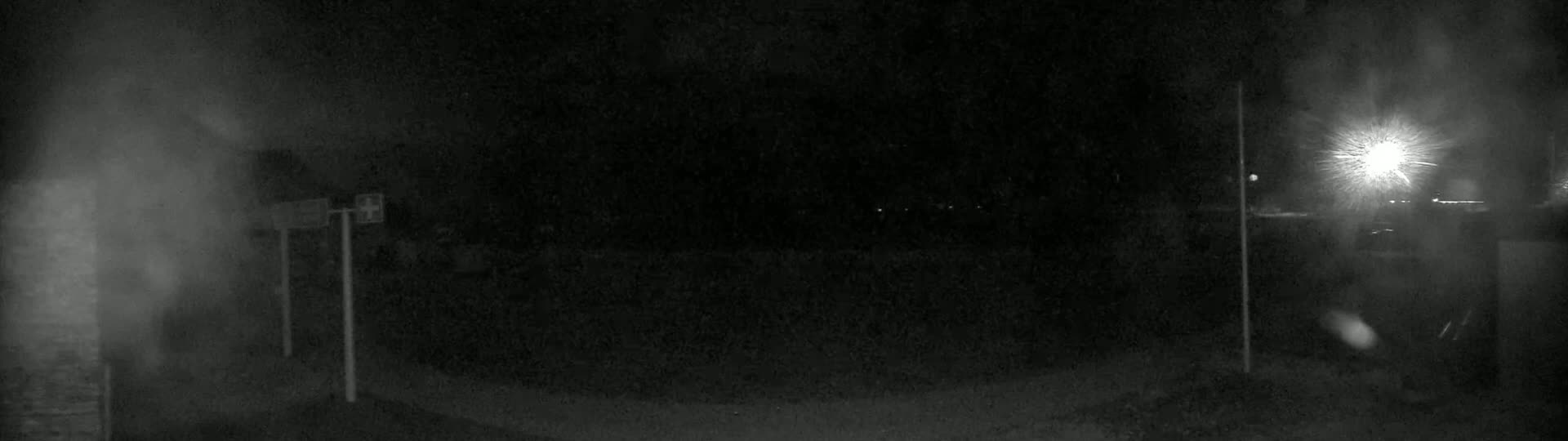 A dark, nighttime scene shows a blurry, poorly lit area with two posts and a bright light source in the distance.