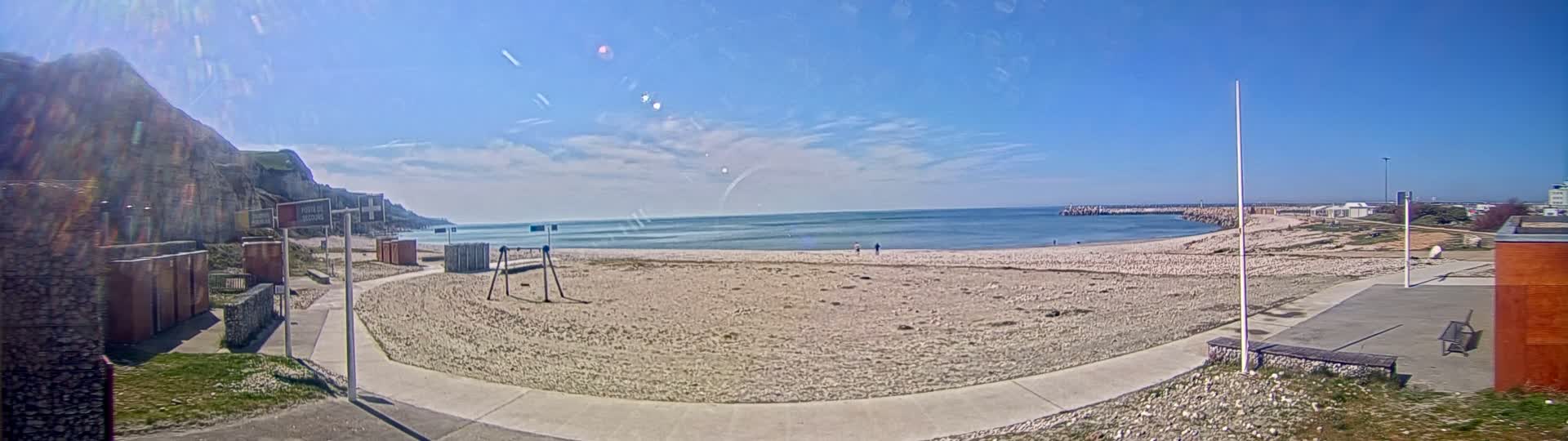 A sandy beach curves gently around a calm, blue ocean under a mostly sunny sky, backed by a tall cliff and a few buildings.