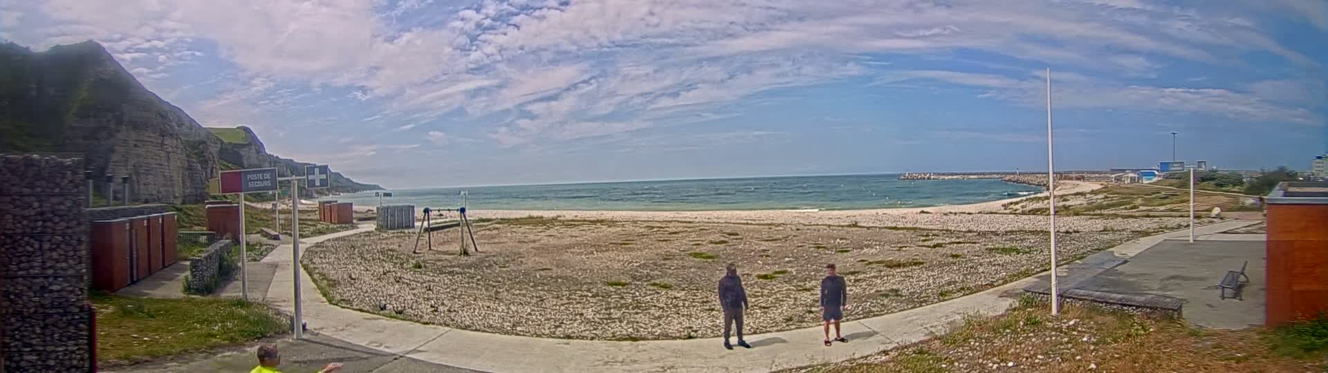 A mostly sunny day at a beach with calm ocean waves, featuring a rocky shoreline, two people standing on a paved path near a sparsely vegetated beach, and a building in the background.