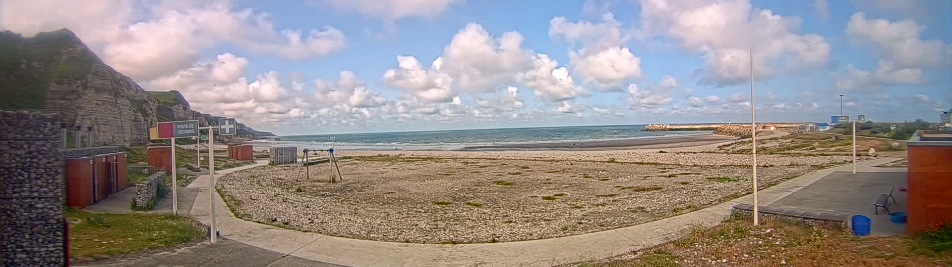 A mostly sunny day at a beach with a pebble shore, showing a small harbor in the distance and a rocky cliff to the left, with some sparse vegetation and a paved walkway.
