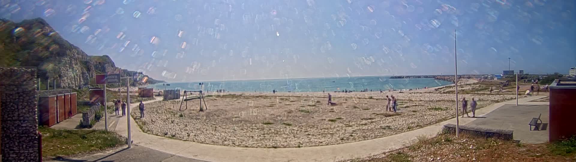 A sandy beach with a few people on it stretches along a calm, light-blue sea under a mostly sunny sky speckled with what looks like lens flares or water droplets.