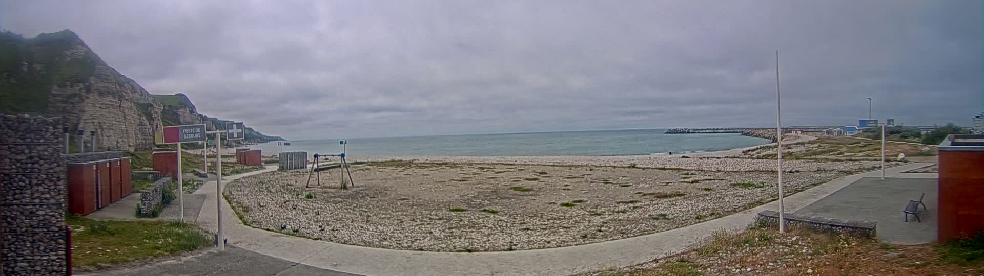 A mostly cloudy day reveals a pebble beach, a small playground, and a rocky cliff overlooking a calm sea, with a few buildings visible in the distance.