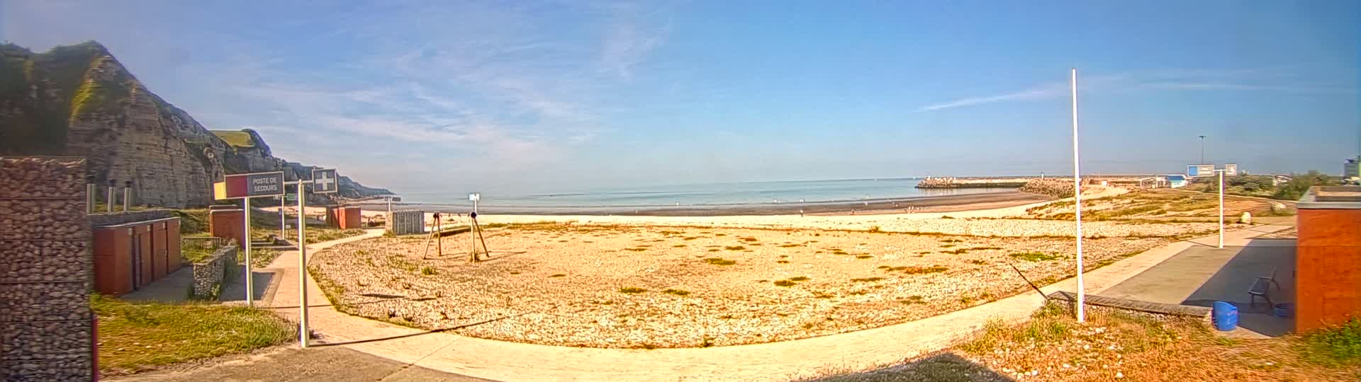 A sandy beach with a low, rocky cliff in the background on a sunny day, near a harbor breakwater.