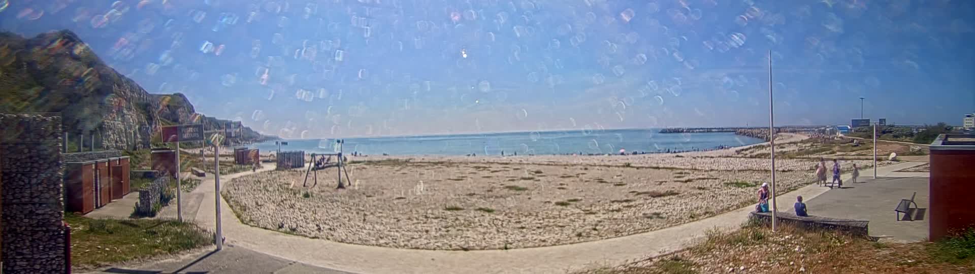 A sandy beach with a few people on it, under a clear blue sky with a hazy overlay, is bordered by a paved path and stone structures, and backed by a cliff.