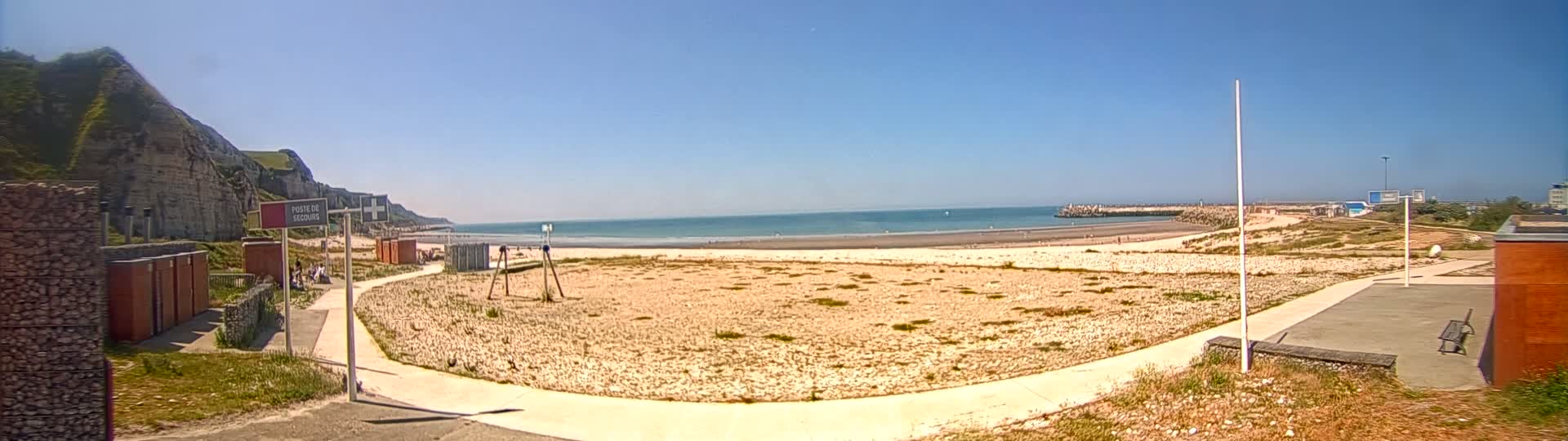 A sandy beach stretches out to a calm ocean under a clear blue sky, backed by a rocky cliff and a man-made breakwater.