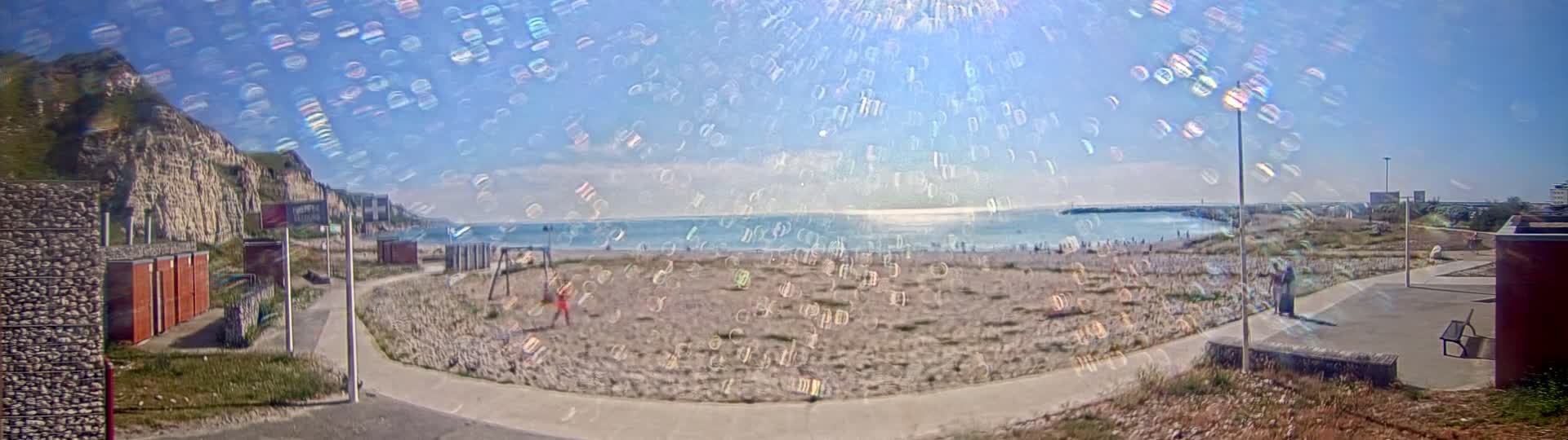 A sandy beach with a few people on it is situated below a cliff face, all under a mostly sunny sky with iridescent light effects.