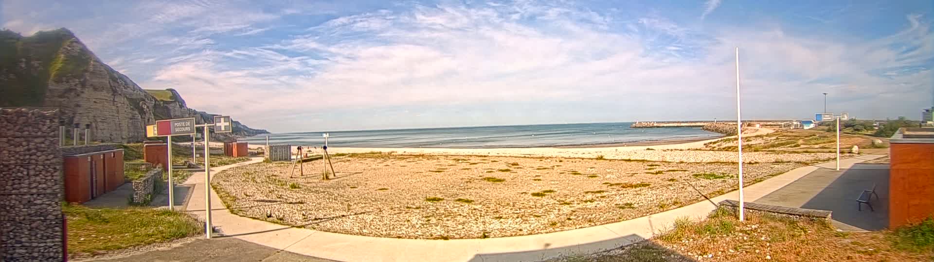 A mostly sunny day at a beach with calm water, featuring a sandy, sparsely vegetated area, a walkway, and white cliffs in the background.