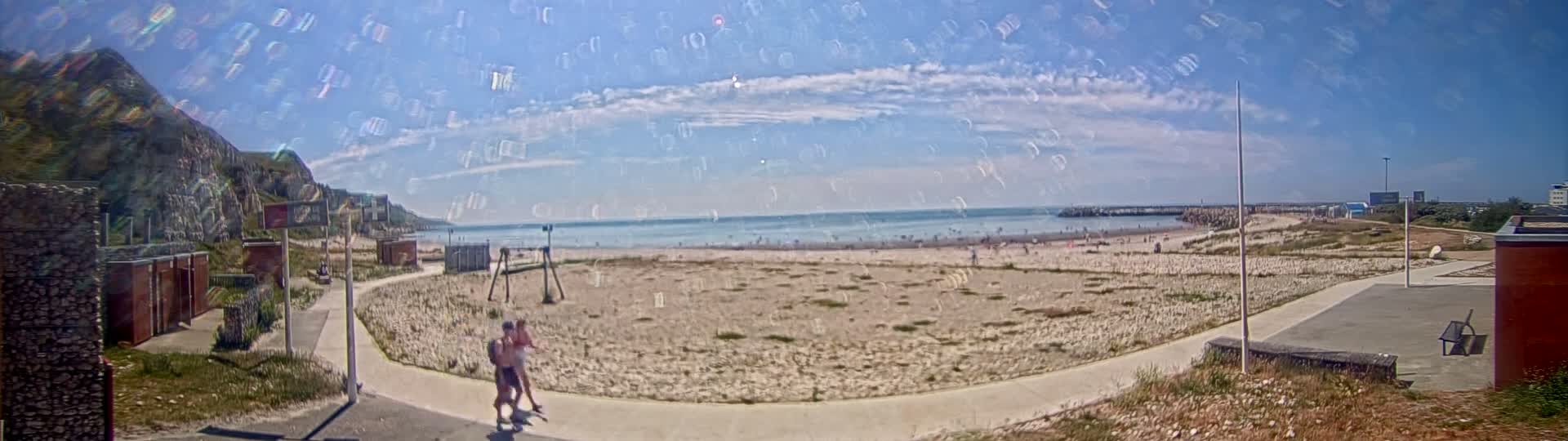 A mostly sunny beach scene shows a sandy expanse with a few people, a walkway, and a rocky cliff in the background.