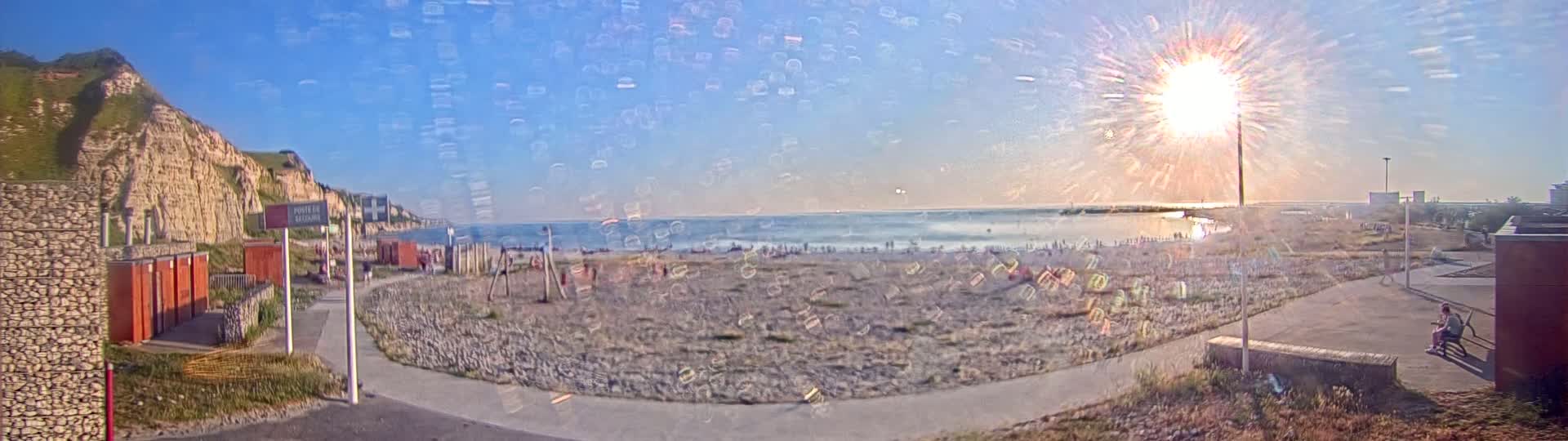 A sandy beach with people on it is situated below a tall, grassy cliff face, all under a bright, sunny sky.