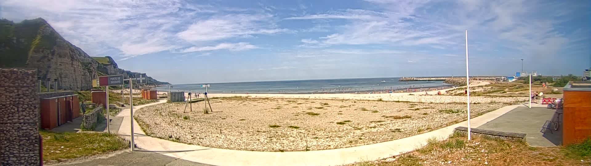 A sandy beach with a few people on it stretches out to a calm ocean under a partly cloudy sky, backed by a rocky cliff and a breakwater.